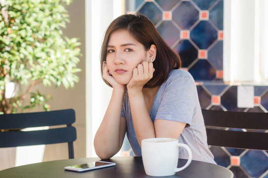 Cheerful asian young woman sitting in cafe drinking coffee and using smartphone for talking, reading and texting. Attractive asian woman holding a cup of coffee while looking on her phone.