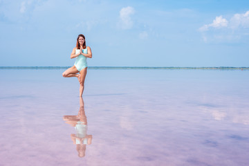 A beautiful woman in a mint body stands in a tree position in a pink salt lake. Redhead young woman doing yoga standing ankle-deep in water, pink salt lakes Bursol'