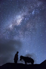Fototapeta premium Horsemen standing under million stars in sky with his horse near smoking hot volcano Mount bromo at night, Indonesia