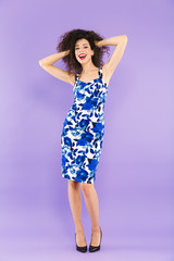 Fototapeta na wymiar Full length photo of happy adorable woman in dress smiling and touching curly hair, isolated over violet background in studio