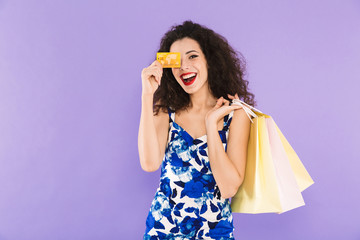 Portrait of beautiful woman 20s wearing dress holding credit card and paper shopping bags, standing isolated over violet background