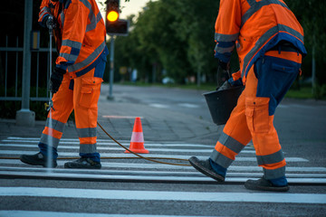 Traffic line painting. Workers are painting white street lines on pedestrian crossing. Road cones with orange and white stripes in background, standing on asphalt during road construction works