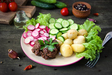 Home made meatballs are served with young potatoes, salad, sliced radishes and cucumbers. In a plate on a wooden table.