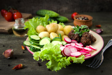 Home made meatballs are served with young potatoes, salad, sliced radishes and cucumbers. In a plate on a wooden table.