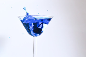 Cocktail with blue liquid in glass. Glass with blue water pouring with liquid with splashes and drops. Martini glass filling with alcohol with splashes on white background. Refreshing drink concept.