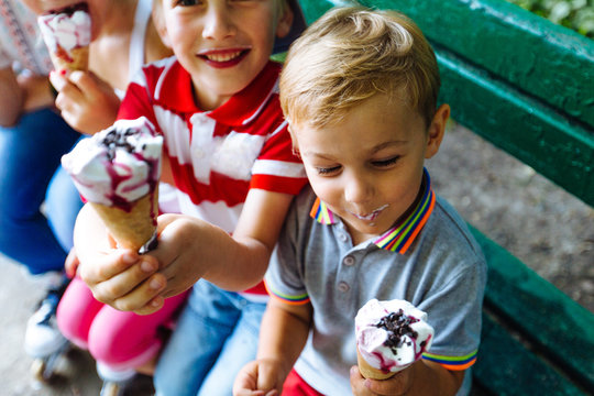 Happy children eating ice cream together outdoor. Photo of happy blond girls with two handsome boys sitting on the bench and smiling at camera.Sun glare effect
