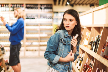 Young frightened woman in denim jacket trying steal bottle of wine in modern supermarket - obrazy, fototapety, plakaty