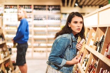 Young serious woman in denim jacket frightenedly looking aside while trying steal bottle of wine in modern supermarket - obrazy, fototapety, plakaty
