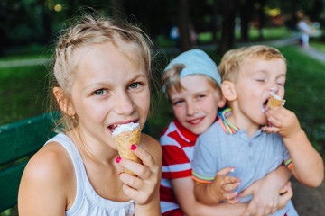 Team of three different age happy children eating ice-cream and having fun together outdoors in...