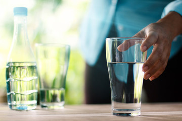 Hand touching and reaching for the glass of drinking water on wooden table, green nature background. Healthy concept