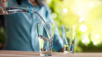 Woman's hand pouring drinking water from bottle to the glass on wooden table, green nature...