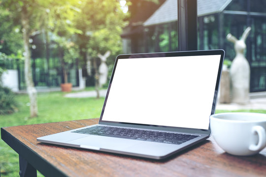 Mockup image of laptop with blank white desktop screen and coffee cup on wooden table with nature background