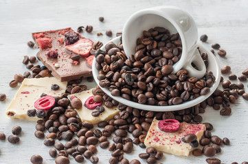 Grains of coffee falling out of a cup and pieces of white chocolate with fruit on a light wooden rough background
