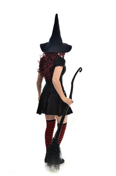 full length portrait of red haired girl wearing black witch costume and pointy hat, holding a broom. standing pose, isolated on white studio background.