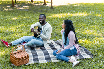 smiling african american boyfriend playing acoustic guitar for girlfriend in park
