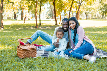 smiling african american parents and daughter sitting at picnic and looking at camera in park