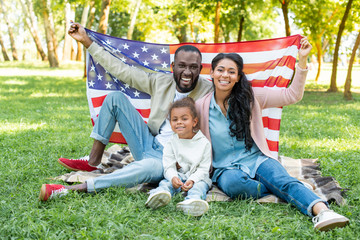 smiling african american parents and daughter holding american flag at picnic in park