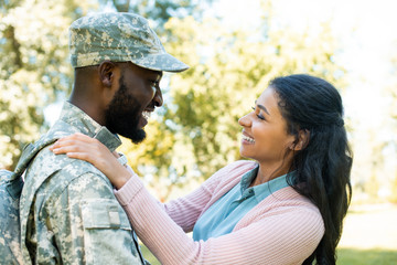 Side view of smiling African American soldier in military uniform hugging girlfriend in park