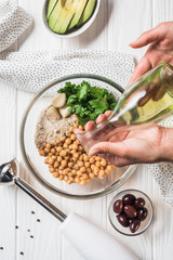 Cropped shot of woman pouring olive oil into bowl with chickpeas and other ingredients for hummus on wooden tabletop