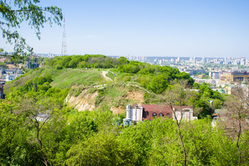 Fototapeta na wymiar Panoramic view of the Zamkova Hora (Castle Hill or mount) in Kyiv covered with greenery. In the background cityscape of Podil and Obolon districts against the blue sky