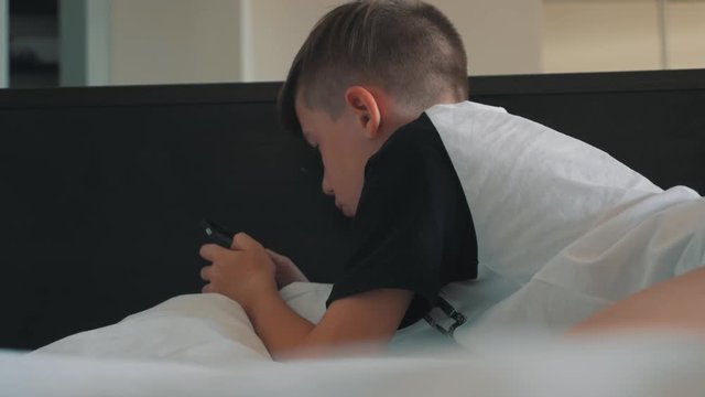 A boy with a smartphone is lying on the bed. The boy communicates with friends with the help of a smartphone
