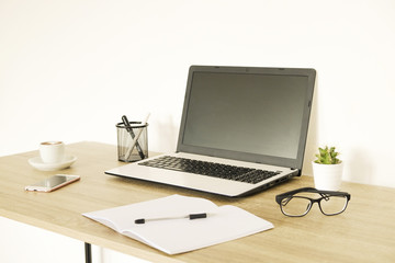 Blank screen laptop computer, cup of cappuccino coffee, cactus, supplies and folded eye glasses on wooden desk in spacious office full of sunlight. Creative workspace. Close up, copy space, background