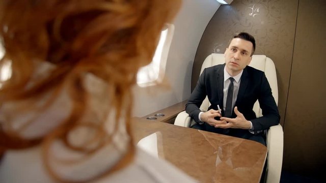 Male chief and female crypto busines partners talk discussing their strategy in the plane. Businessman in black suit and lawyer businesswoman with red hait sitting opposite to each other at the table