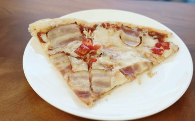 Close-up of pizza slice with bacon and sweet pepper on white plate on wooden table.