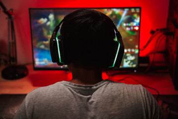 Portrait from back of teenage gamer boy looking at screen and playing video games on computer in...