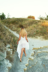 Girl goes on coastal desert path, with dried grass. Blonde in a white tunic. The theme of the desert and the sea