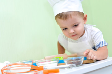 A cute two year old boy dressed as a chef makes a cookie, looks in a bowl with raisins and wants to take it for a biscuit or taste.