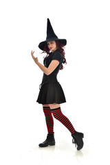 full length portrait of red haired girl wearing black witch costume and pointy hat.  standing pose, isolated on white studio background.