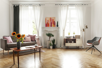Sunflowers on wooden table next to sofa in spacious flat interior with armchair and poster. Real photo - obrazy, fototapety, plakaty
