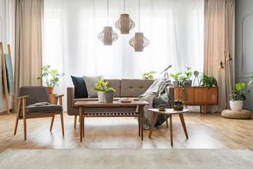 Lamps above wooden table with flowers in bright living room interior with sofa and armchair. Real...