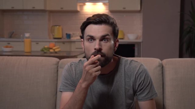 Guy sits on sofa and watches football game. He eats popcorn. Suddenly his team loses. Guy looks dissapointed. He leans forward and backwards and holding hands on head. Then man continue to watch.