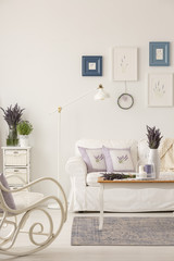 Metal lamp placed by the sofa in real photo of white living room interior in provencal style with fresh lavender and posters on wall