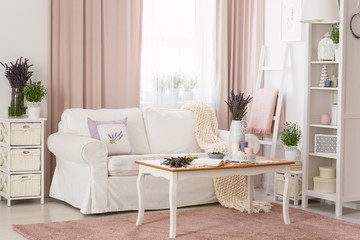 Real photo of bright provencal sitting room interior with white sofa, wooden coffee table on dirty pink carpet, rack with decor and fresh lavender - obrazy, fototapety, plakaty