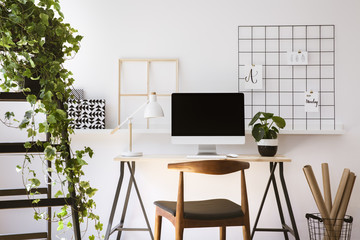 Real photo of wooden desk with metal lamp, fresh plant and empty screen monitor standing in white flat interior