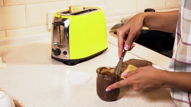 Camera is focused on girl's hands. She takes one toast from toaster. Then girl takes nife and get some nutella out of jar and out it on fried bread. She lubricates it on toast.