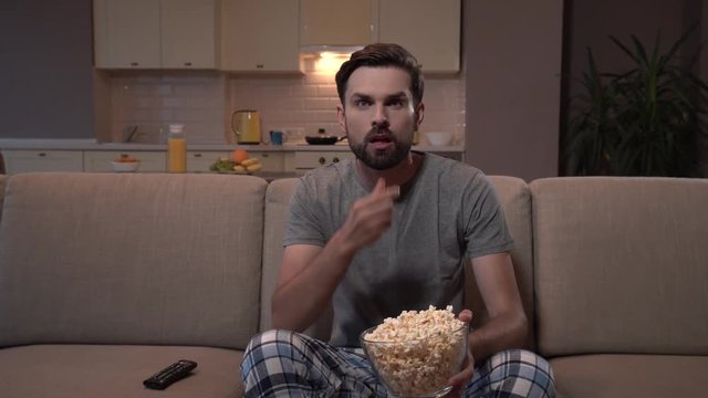 Guy sits on sofa and watches TV. He eats popcorn. Guy hs it in glass bowl. He is concentrated on watching. He looks straight forward.