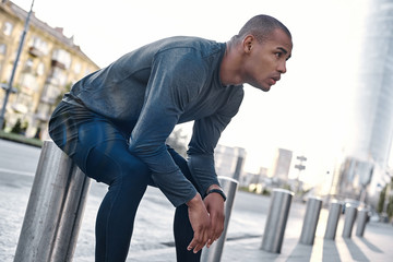 Urban sports. Young man runner resting after workout session on sunny morning. Male fitness model sitting on street along pond in city