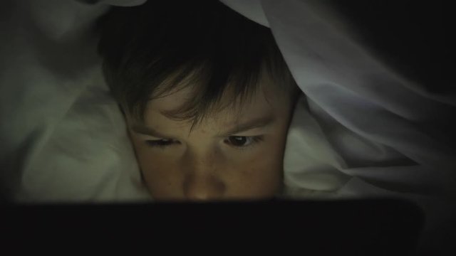 A happy boy lies in bed under a blanket and plays on a tablet in a game in the dark. The face of the child is lit by a bright monitor