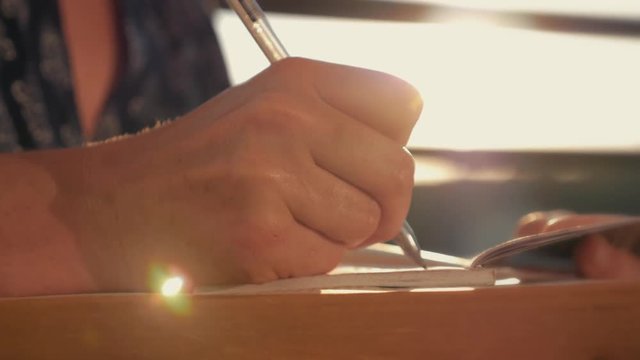 Woman's hand close-up, writing a note or letter to a notebook with a pen on a sunny day at sunset. A stack of scientific books, preparation for exams. Filling out the report