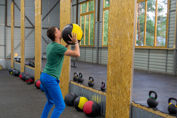 guy in a green T-shirt and blue pants trains in the gym, throws a medical ball against the wall, on the background of rows of weights with multi-colored handles and a composition of sports equipment