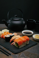 food composition with sushi set, ginger and soya sauce in bowls, teapot and cups with tea on dark surface