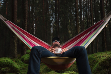A man with a mug of tea or coffee sitting on a hammock in a forest