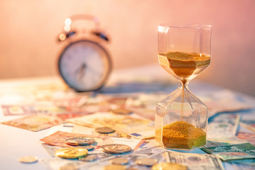 Obraz na płótnie Canvas Sand running through the shape of hourglass on table with banknotes and coins of international currency. Time investment and retirement saving. Urgency countdown timer for business deadline concept
