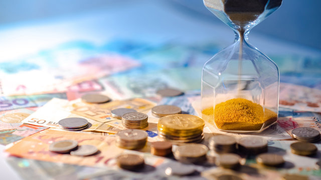 Sand running through the shape of hourglass on table with banknotes and coins of international currency. Time investment and retirement saving. Urgency countdown timer for business deadline concept