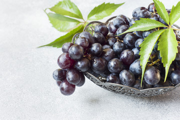 Fresh black grapes with leaves in a plate on a grey table selective focus. Copy space