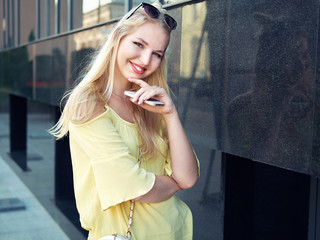 Young beautiful blonde woman with multi colored eyes high bun hairstyle jeans shorts yellow blouse enjoying warm evening posing against city building wall
